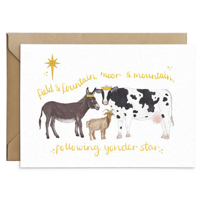 A unique christmas card lays on brown envelope on a white background. The card depicts a whimsical illustration farm animals as the three wise men- a donkey, goat and cow.  Hand scripted yellow text reads 'field and fountain, moor and mountain, following yonder star'.