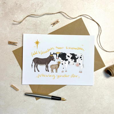 A unique christmas card lays on a table on top of a brown envelop beside a fountain pen. The card depicts a whimsical illustration farm animals as the three wise men- a donkey, goat and cow.  Hand scripted yellow text reads 'field and fountain, moor and mountain, following yonder star'.