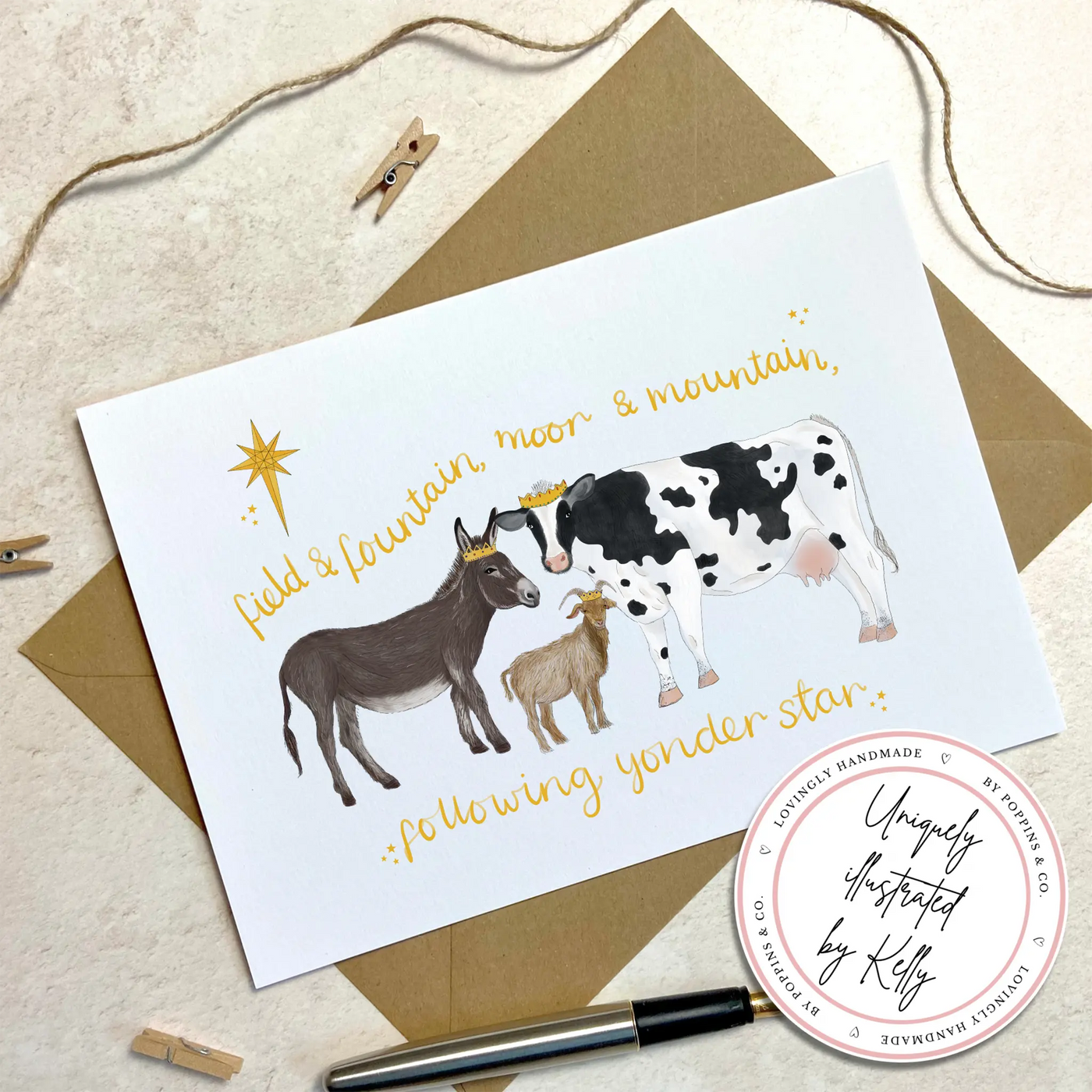 A unique christmas card lays on a table on top of a brown envelope, beside a fountain pen. The card depicts a whimsical illustration farm animals as the three wise men- a donkey, goat and cow.  Hand scripted yellow text reads 'field and fountain, moor and mountain, following yonder star'.