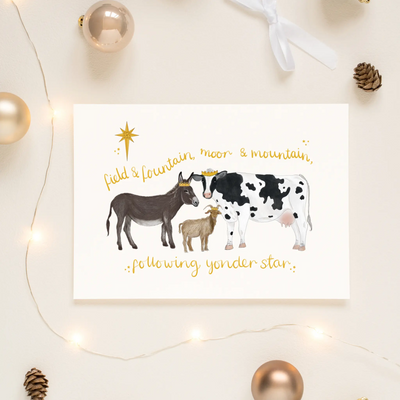 A unique christmas card lays on a table surrounded by christmas lights and ornaments. The card depicts a whimsical illustration of a donkey, cow and goat wearing golden crowns beneath the star of Bethlehem. Hand scripted yellow text read 'field & fountain, moor & mountain, following yonder star.'