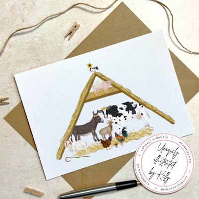 A unique christmas card lays on a table surrounded by pegs and a pen. The card depicts a whimsical illustration of nativity scene where all the roles are played by farm yard animals. 