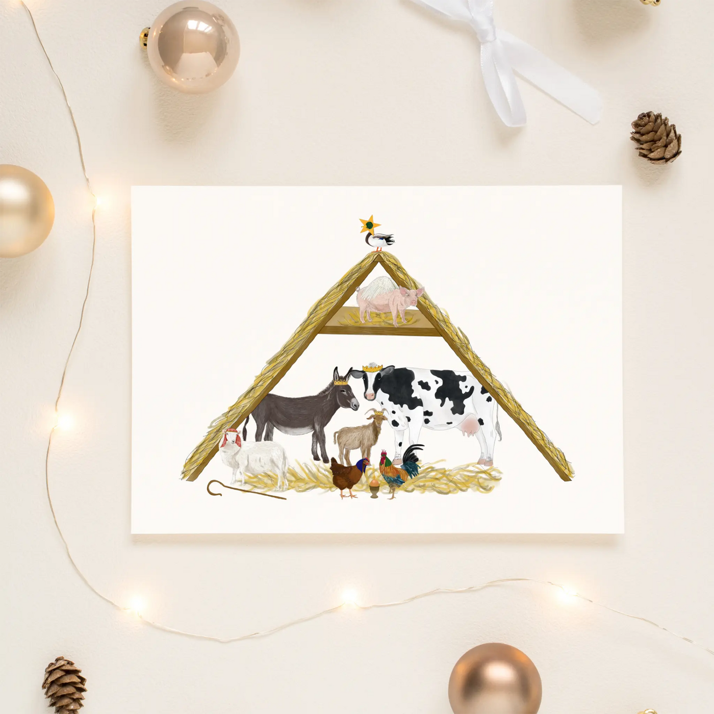 A unique christmas card lays on a table surrounded by christmas lights and ornaments. The card depicts a whimsical illustration of a variety of farm animals taking the roles of traditional Nativity characters in this traditional nativity stable scene. A chicken, cockerel and egg represent Mary, Joseph and Baby Jesus, a donkey, cow and goat are the three kings and the scene is completed by a sheep shepherd, angel pig and little duck as the star of Bethlehem.