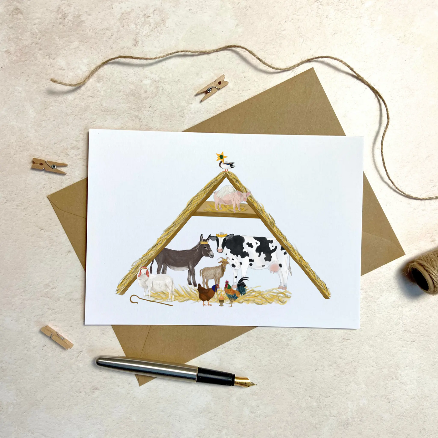 A unique christmas card lays on a table surrounded by christmas lights and ornaments. The card depicts a whimsical illustration of nativity scene where all the roles are played by farm yard animals. 