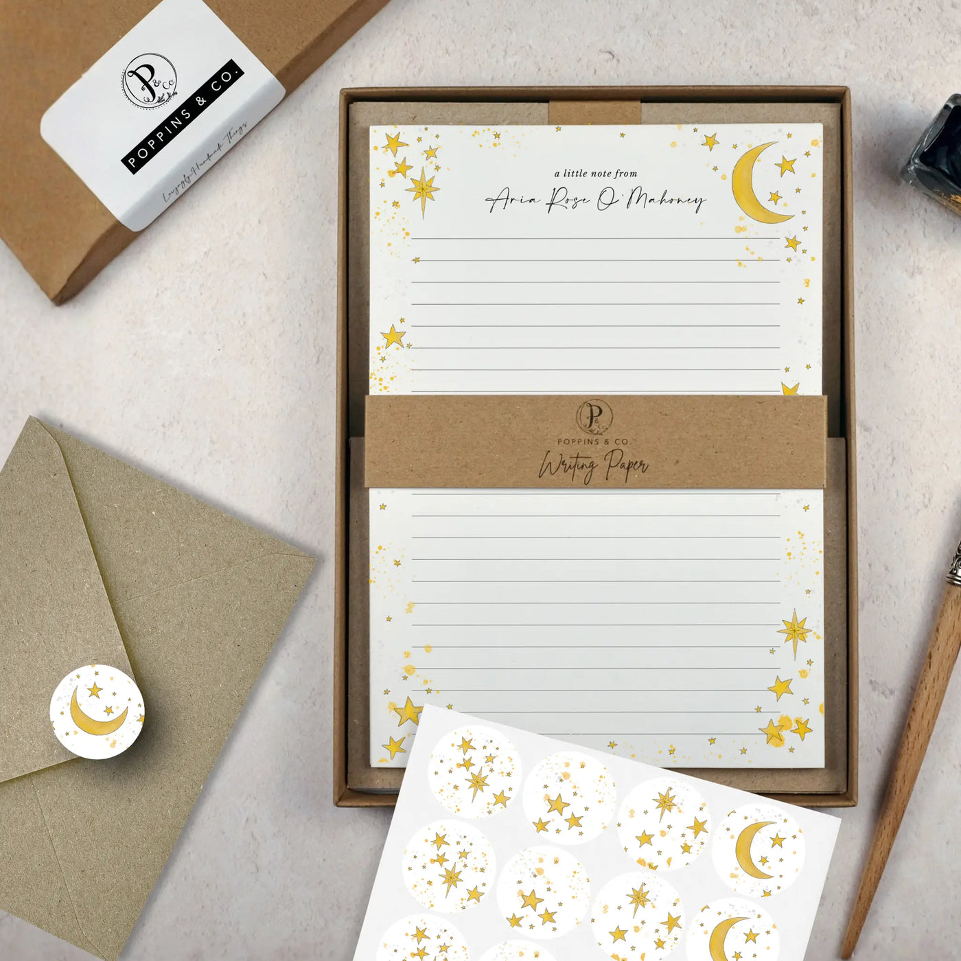 A celestial themed writing set by in a brown kraft luxury gift box. The writing paper is white and has illustrations of a yellow stars and moons around the border. Italic script reads 'a little note from' with the name 'Aria Rose O'Mahoney' typed in in a script font. On top of the box lays a sheet of round envelope stickers with the star and moon designs. To the left lays a brown kraft envelope with an envelope sticker seal on it.