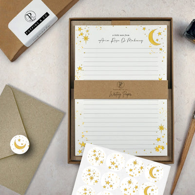 A celestial themed writing set by in a brown kraft luxury gift box. The writing paper is white and has illustrations of a yellow stars and moons around the border. Italic script reads 'a little note from' with the name 'Aria Rose O'Mahoney' typed in in a script font. On top of the box lays a sheet of round envelope stickers with the star and moon designs. To the left lays a brown kraft envelope with an envelope sticker seal on it.