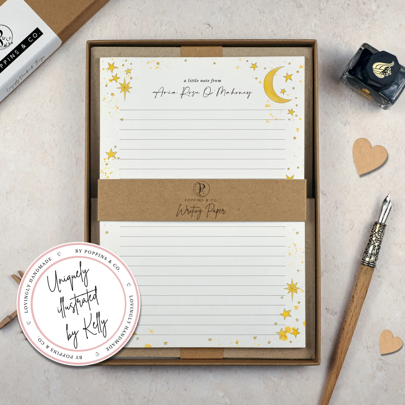 A celestial themed lined writing paper set by in a brown kraft luxury gift box. The writing paper is white and has illustrations of a yellow stars and moons around the border. Italic script reads 'a little note from' with the name 'Aria Rose O'Mahoney' typed in in a script font. 
