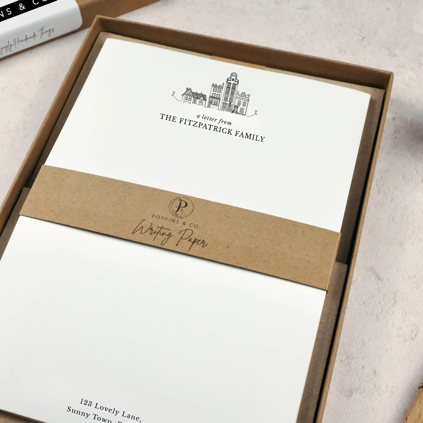 A family writing paper set by in a brown kraft luxury gift box. The writing paper is white and has illustrations of a little line drawn houses in black at the top of the page. Italic script reads 'a letter from' with 'The Fitzpatrick Family' typed in all caps font below it. 