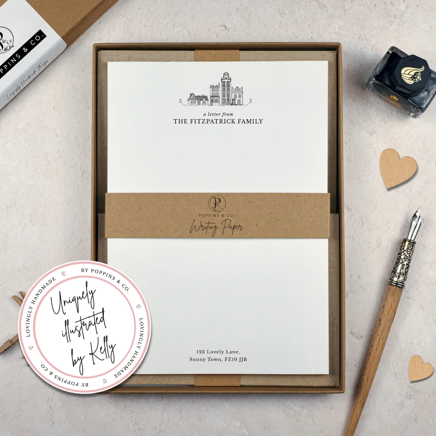 A family writing paper set by in a brown kraft luxury gift box. The writing paper is white and has illustrations of a little line drawn houses in black at the top of the page. Italic script reads 'a letter from' with 'The Fitzpatrick Family' typed in all caps font below it. A sticker on the corner of the photo reads 'uniquely illustrated by Kelly'.