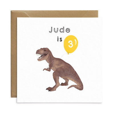 Dinosaur birthday card with unique illustration of a T rex holding a yellow balloon. Personalised text reads 'Jude is' with the number '3' on the balloon. Square card with brown Kraft envelope. Kids Birthday Card by Poppins and co.
