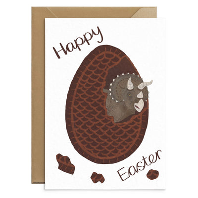 An unusual easter card for kids featuring a brown triceratops hatching from inside chocolate easter egg. Brown text reads 'happy easter'. Illustration on white A6 card with recycled envelopes. Easter cards by Poppins and co.