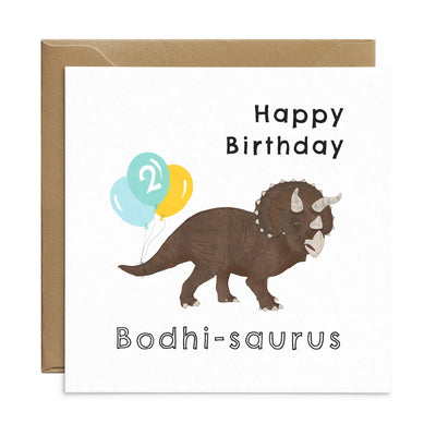 Dinosaur birthday card with unique illustration of a Triceratops with a bunch of balloons tied to its tail. Personalised text reads 'Happy Birthday Bodhi-saurus' with the number '2' on the balloons. Square card with brown Kraft envelope. Kids Birthday Card by Poppins and co.