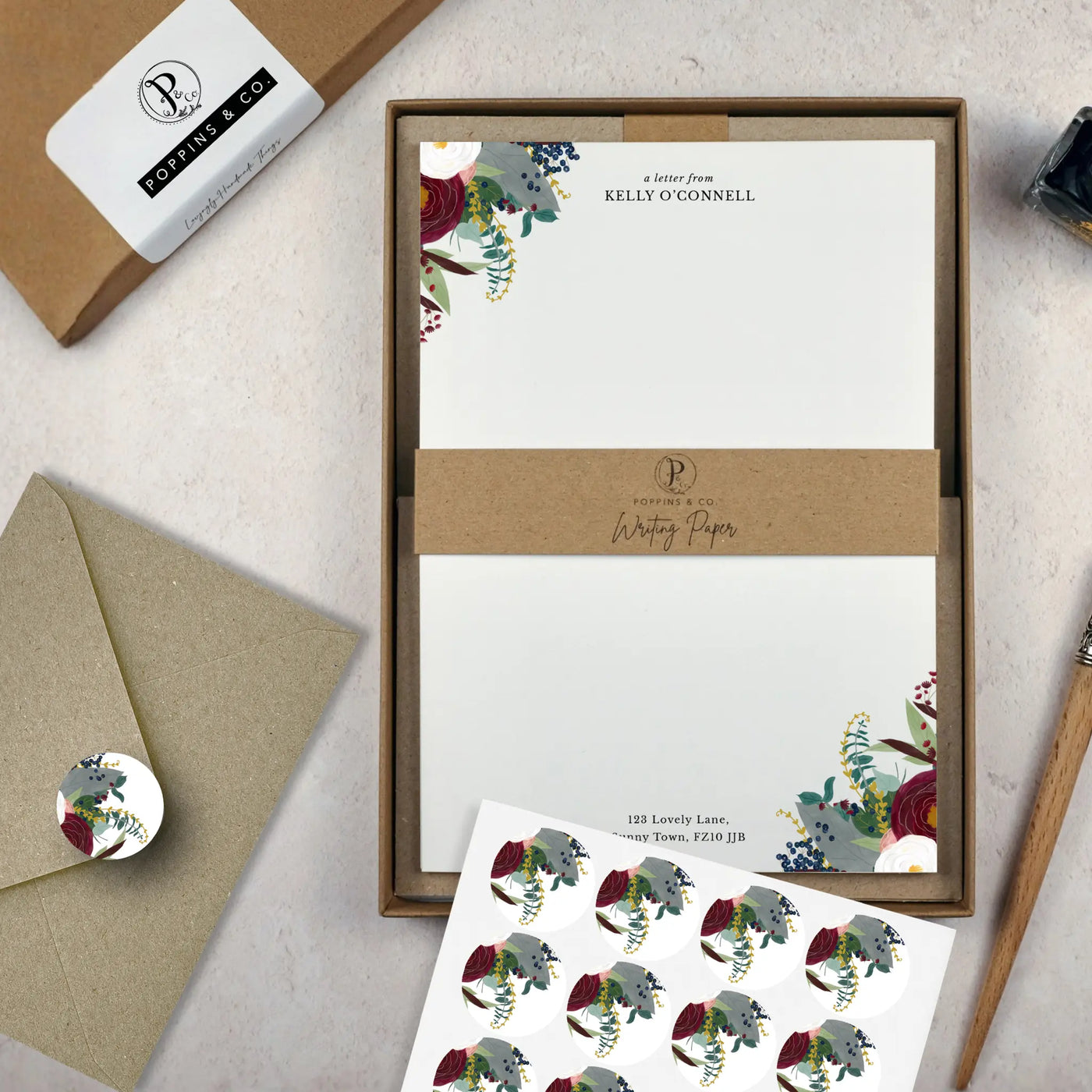 A winter floral writing set by in a brown kraft luxury gift box. The writing paper is white and has illustrations of a burgundy and white flowers and foliage on two corners. Italic script reads 'a letter from' with the name 'Kelly O'Connell' typed in all caps font below it. On top of the box lays a sheet of round envelope stickers with the same floral illustration on each. To the left lays a brown kraft envelope with an envelope sticker seal on it.