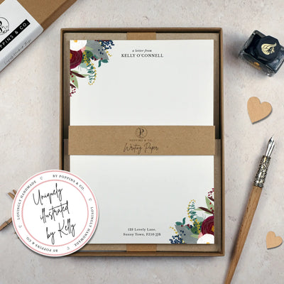 A winter floral writing set by in a brown kraft luxury gift box. The writing paper is white and has illustrations of a burgundy and white flowers and foliage on two corners. Italic script reads 'a letter from' with the name 'Kelly O'Connell' typed in all caps font below it. 