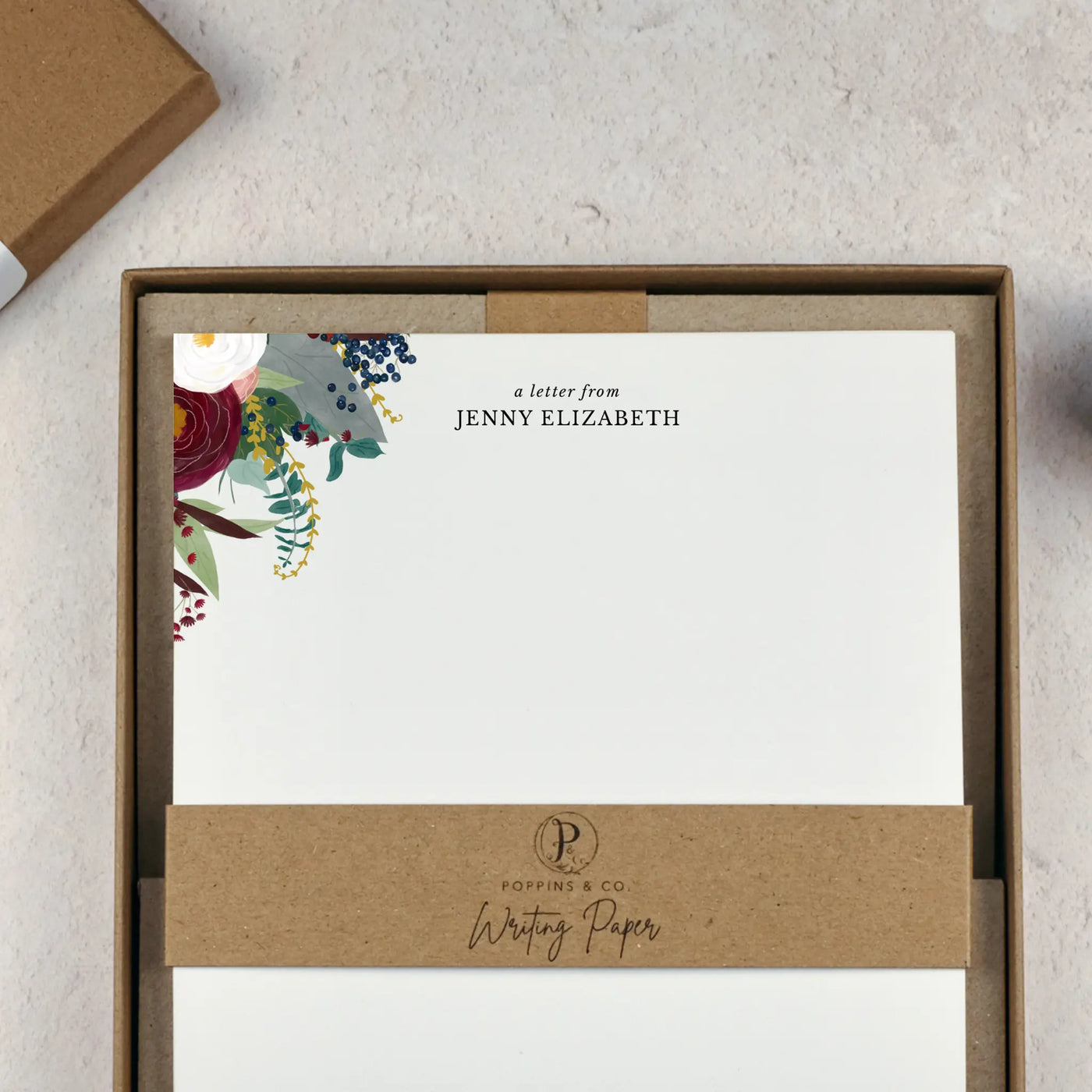 A winter floral writing set by in a brown kraft luxury gift box. The writing paper is white and has illustrations of a burgundy and white flowers and foliage on two corners. Italic script reads 'a letter from' with the name 'Jenny Elizabeth' typed in all caps font below it.